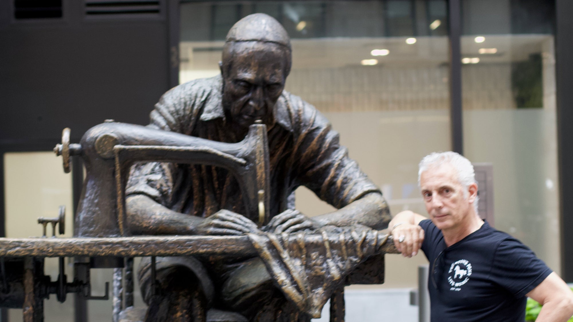 Anthony Lilore posing next to a bronze statue of a person working on a sewing machine in the Garment District NYC