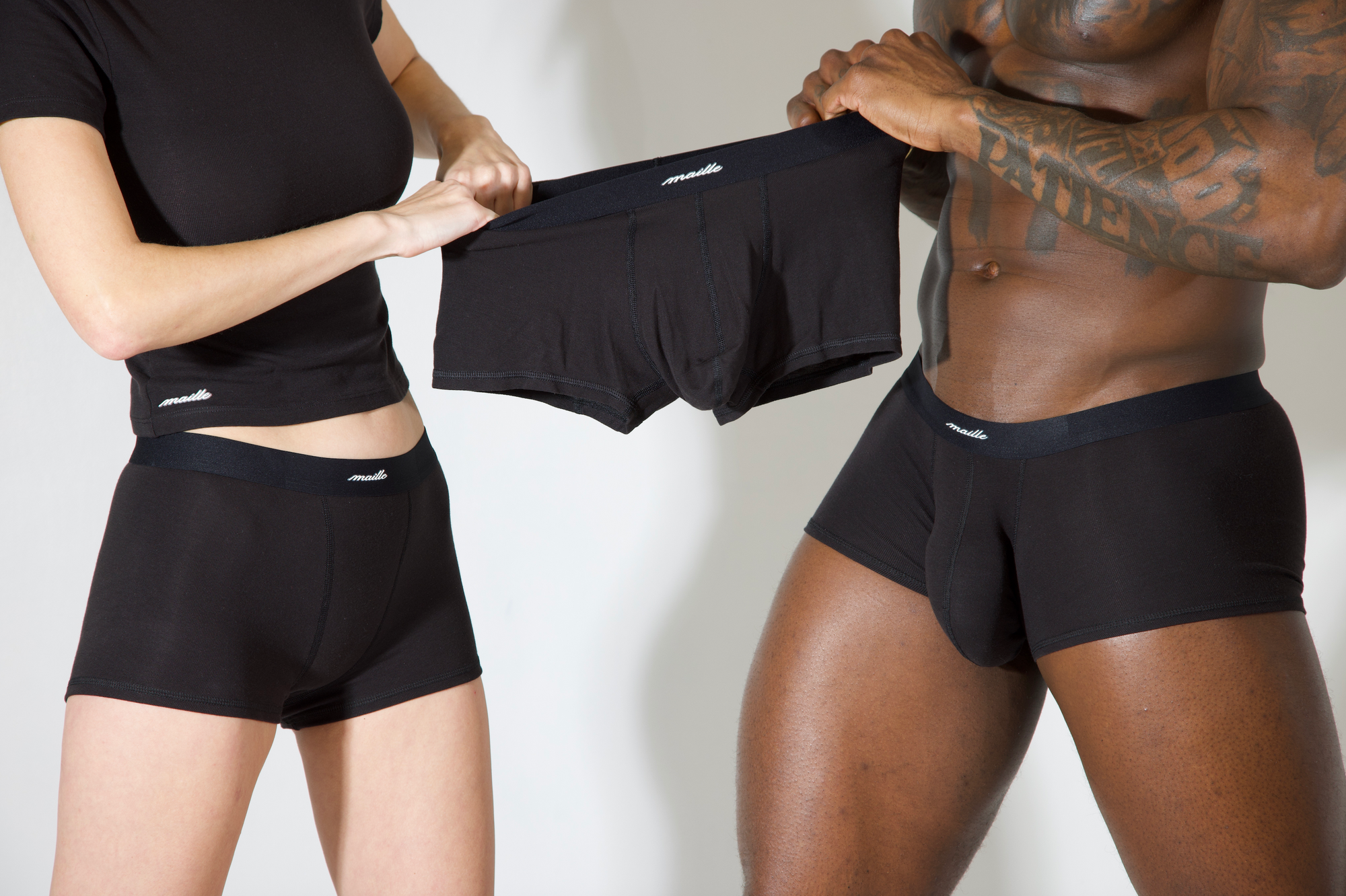 A man and a woman wearing maille underwear in black fighting over a third boxer brief against a white background
